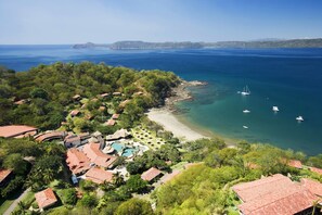 Vista aérea de un complejo turístico costero con varios edificios, una playa y embarcaciones en el agua.