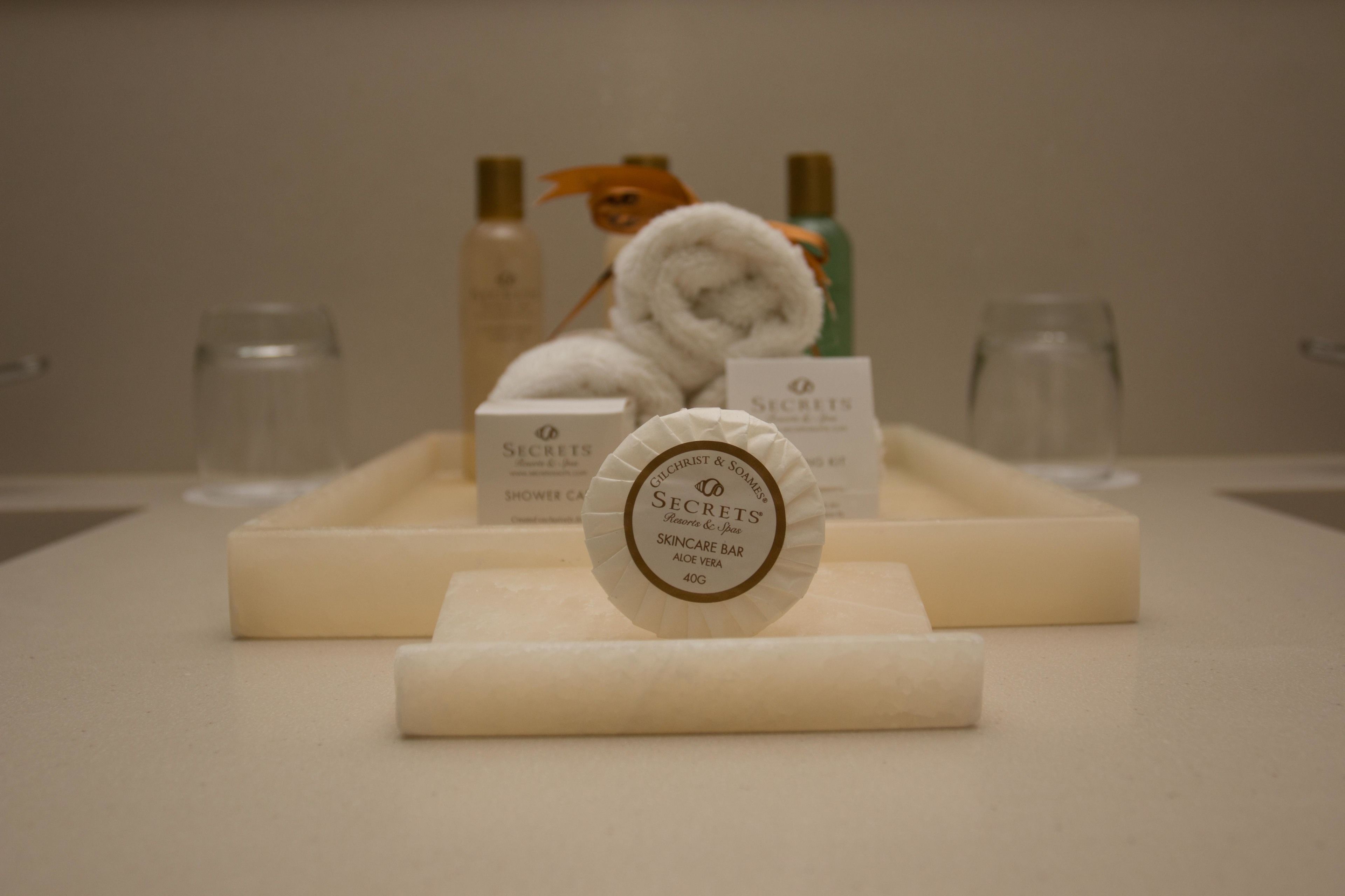 A skincare product display with a white box labeled "Secrets Skincare Bar Aloe Vera 40g" in the foreground, a rolled white towel, and two bottles in the background.