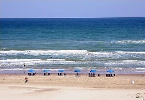 Une plage avec des vagues, une personne qui court et des parasols bleus.