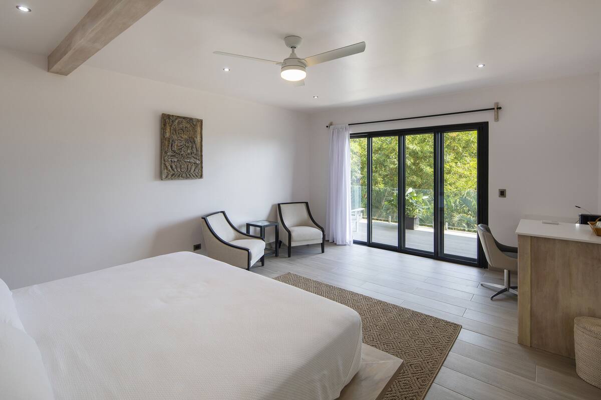 Une chambre d’hôtel moderne dotée d’un grand lit, de deux fauteuils, d’un bureau et d’une porte-fenêtre coulissante donnant sur un balcon.