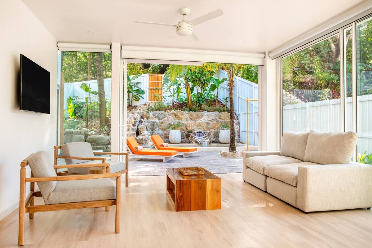 Un vaste espace de vie doté d’un plancher en bois, d’un canapé beige, de deux fauteuils en bois et d’une table basse. Une grande fenêtre offre une vue sur un jardin avec un mur en pierre, des plantes et un élément décoratif aquatique.
