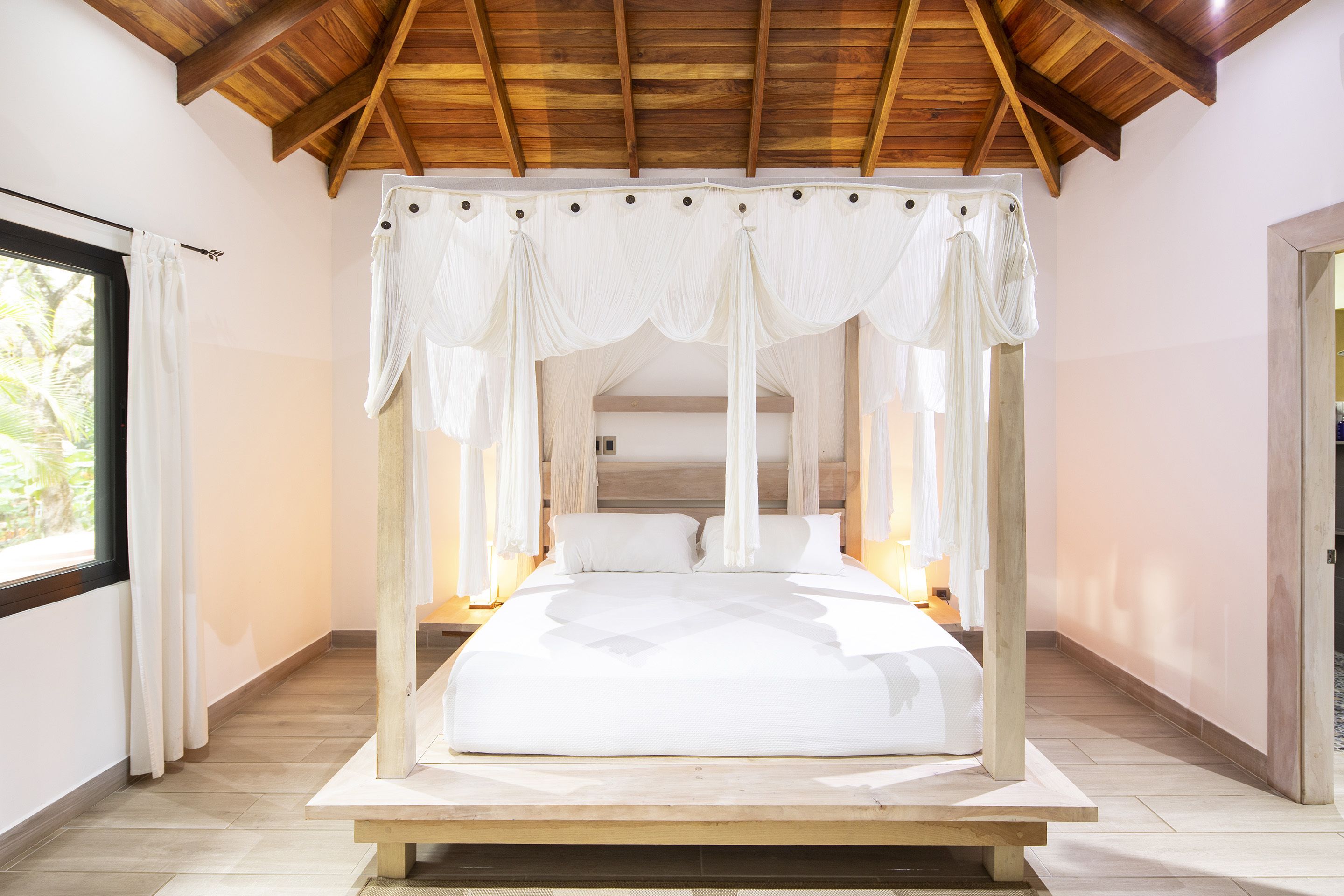 A four-poster canopy bed with white linens, a wooden ceiling, and a view of the outdoors through a window.