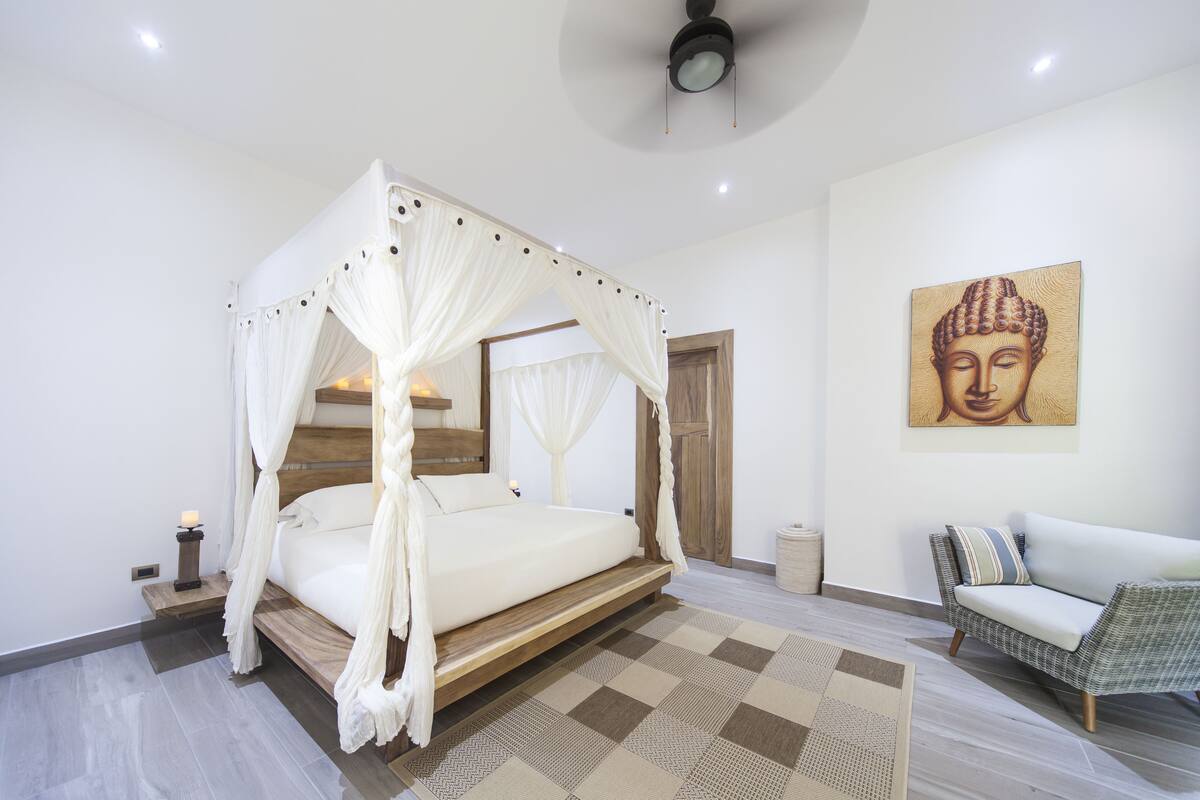 Une chambre à coucher avec un lit à baldaquin, un canapé, un tableau représentant Bouddha et une porte en bois.