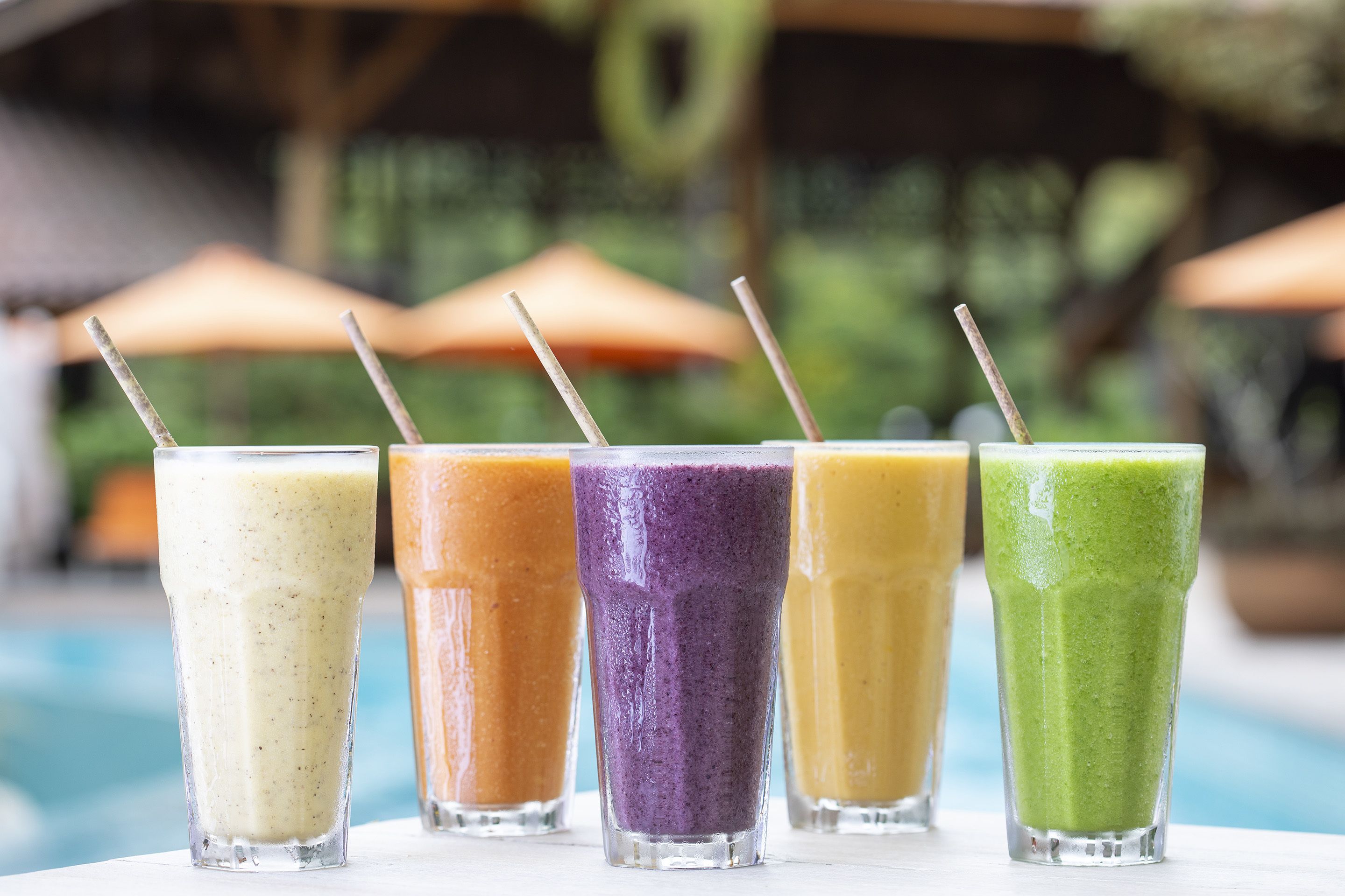 A row of colorful smoothies in clear glasses with straws, placed on a table by a poolside.
