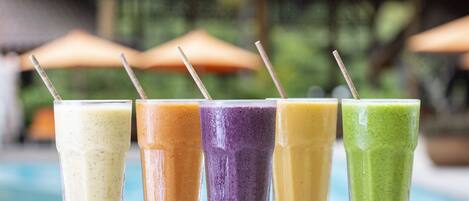A row of colorful smoothies in clear glasses with straws, placed on a table by a poolside.