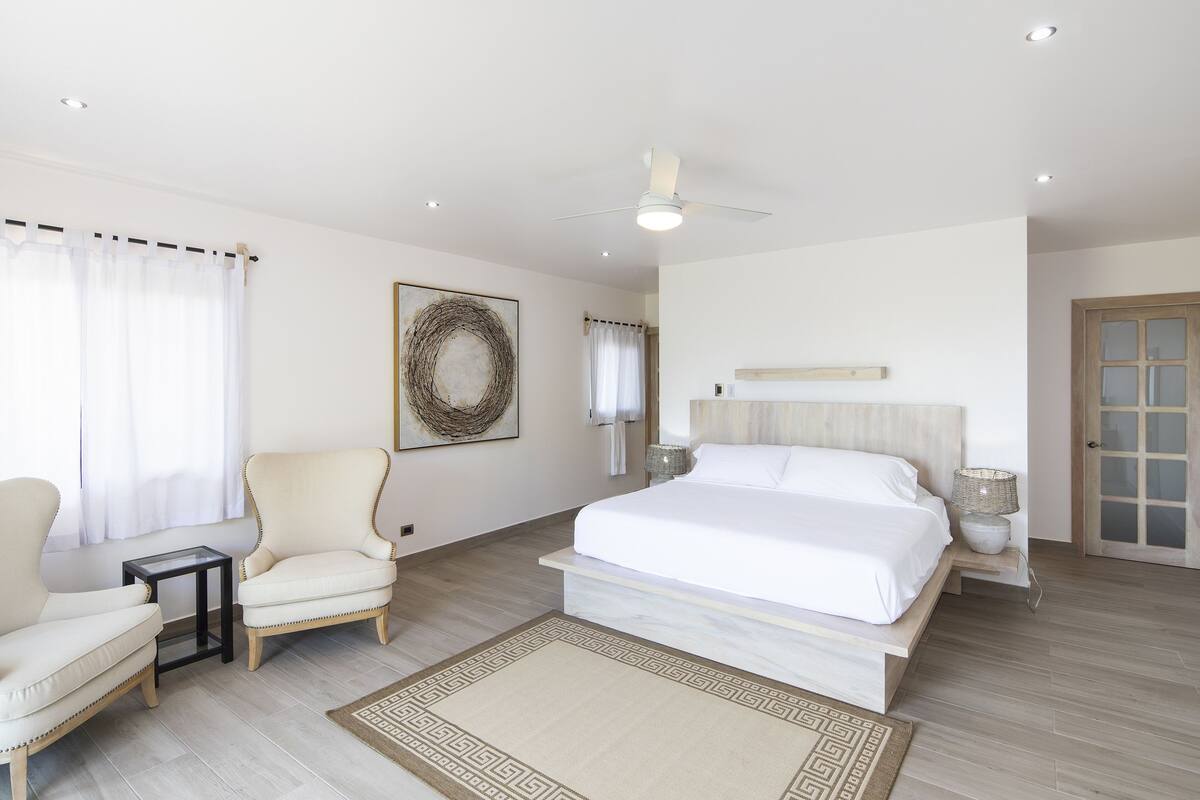 Une chambre spacieuse avec un grand lit, deux fauteuils, une petite table et un ventilateur de plafond.