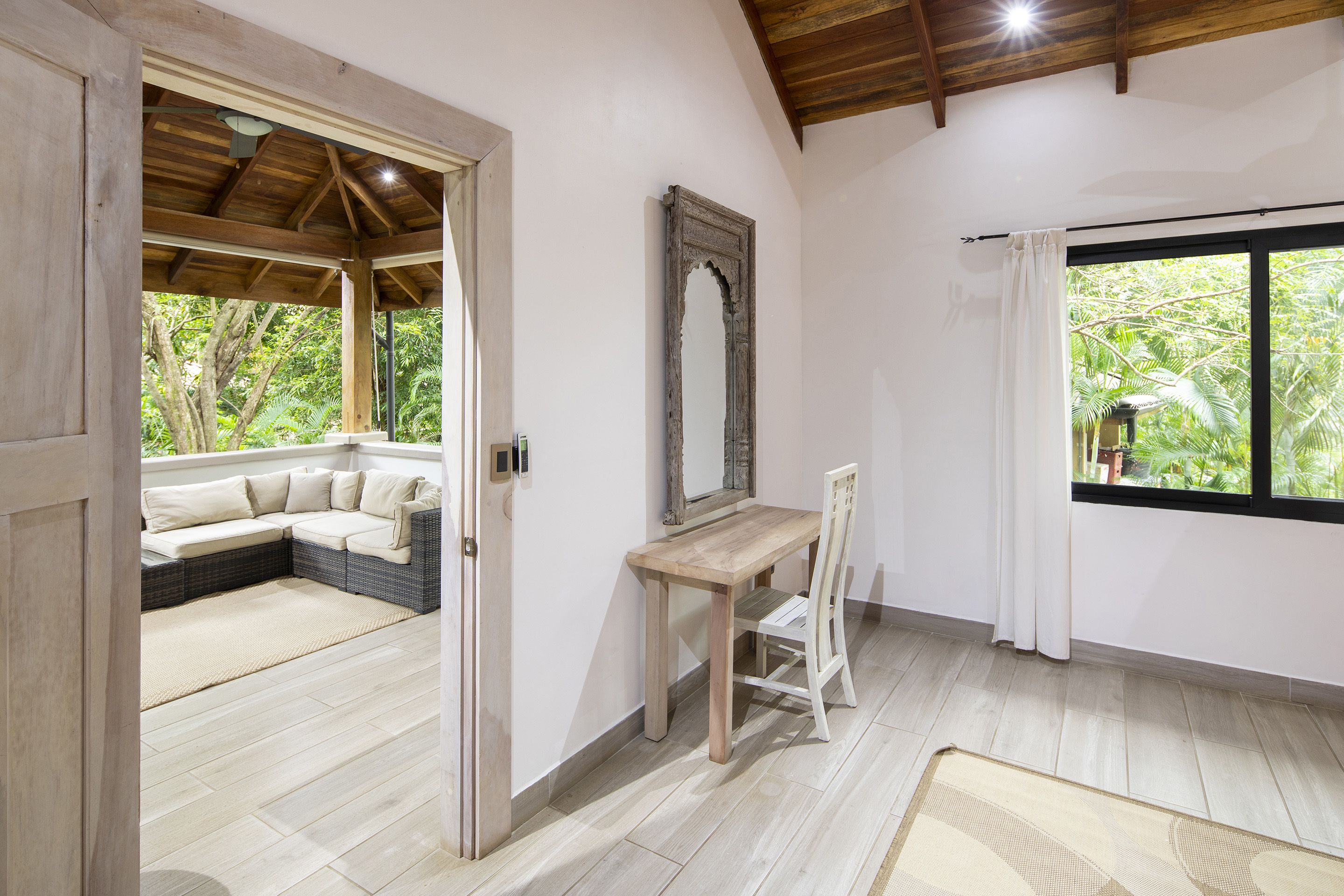 Ein Zimmer mit Holzboden, einem großen Fenster mit Vorhängen, einem Holztisch, einem Stuhl und einer gepolsterten Sitzecke mit Blick auf Grünfläche.