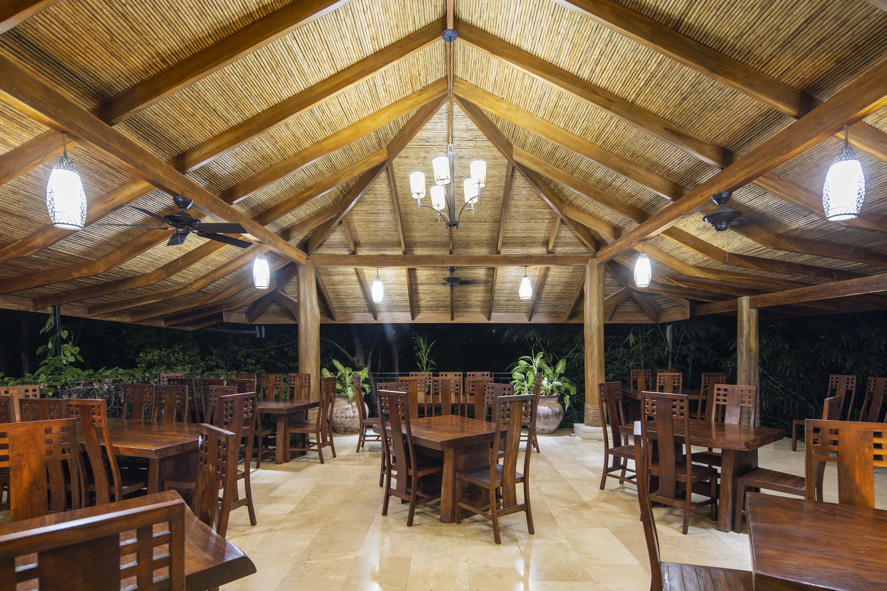An outdoor covered dining area with wooden tables and chairs, a high wooden ceiling, and hanging light fixtures.