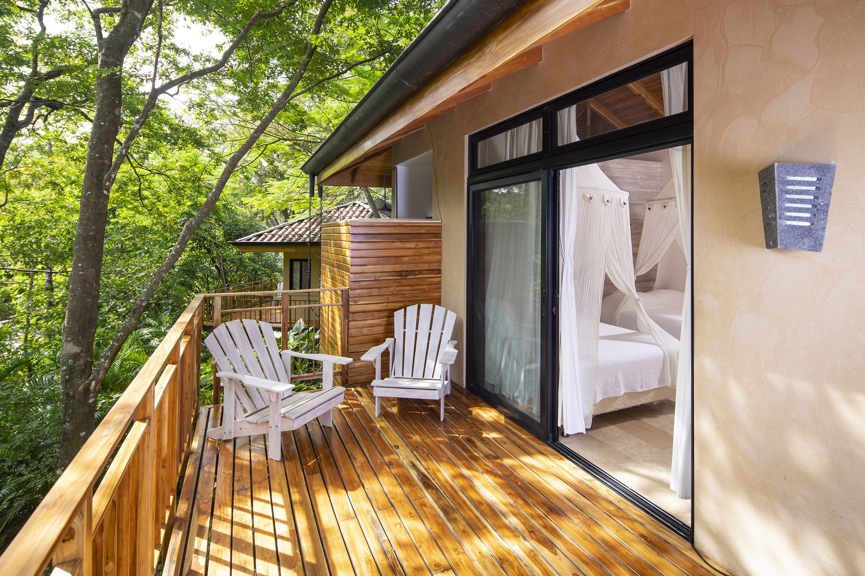 A wooden deck with two white Adirondack chairs and a canopy bed with white curtains.