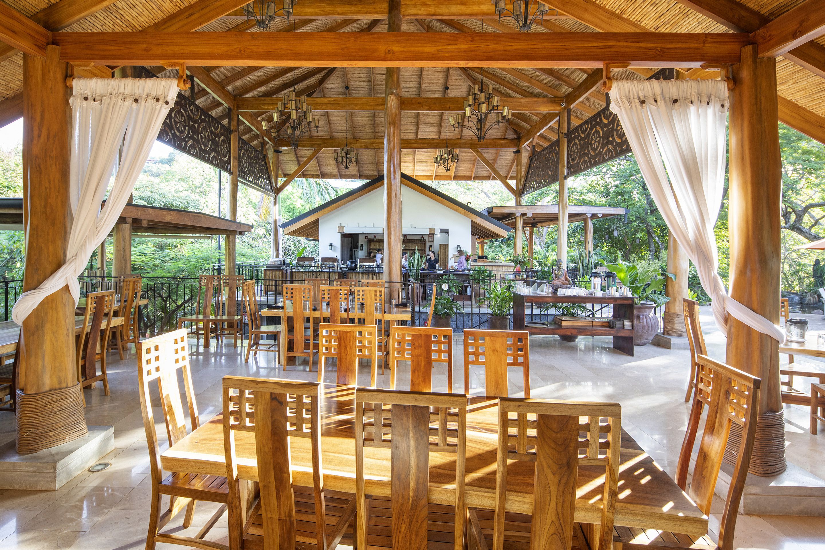 Un espace extérieur aménagé pour les repas, avec des tables et des chaises en bois, un coin salon abrité et une vue sur un bâtiment en arrière-plan.
