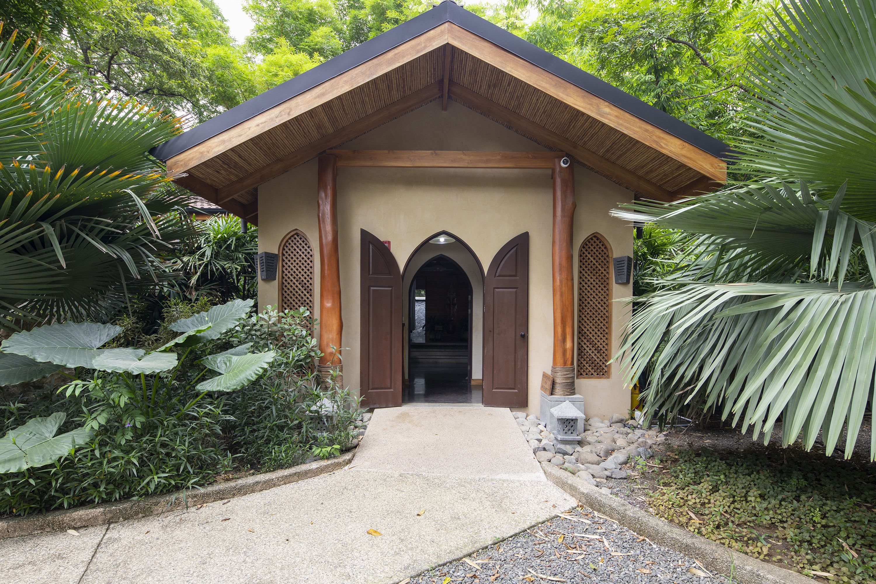 A building with a wooden roof and three arched doorways, surrounded by tropical plants.