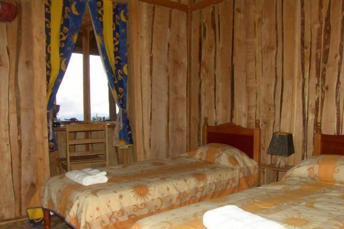 Quarto com duas camas, paredes de madeira, janela com cortinas, mesa de cabeceira e luminária.
