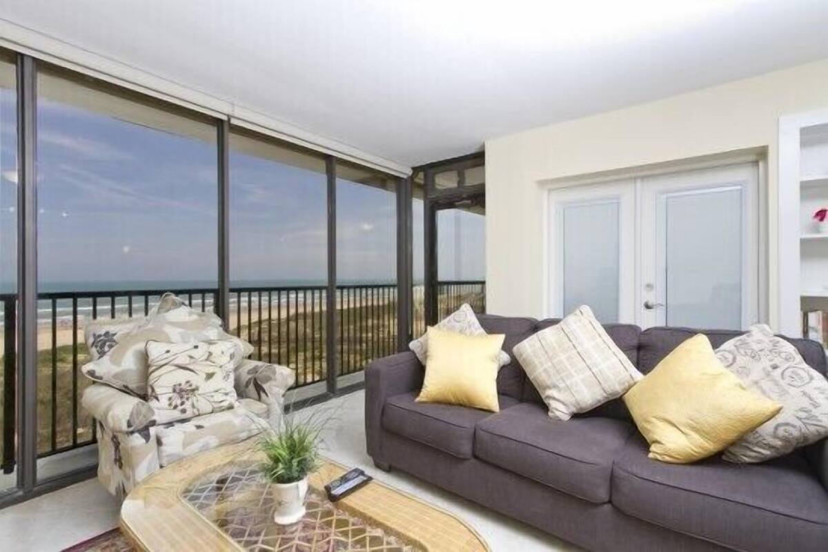 A balcony with a grey sofa and cushions, a small table with a plant, and a view of the sea.
