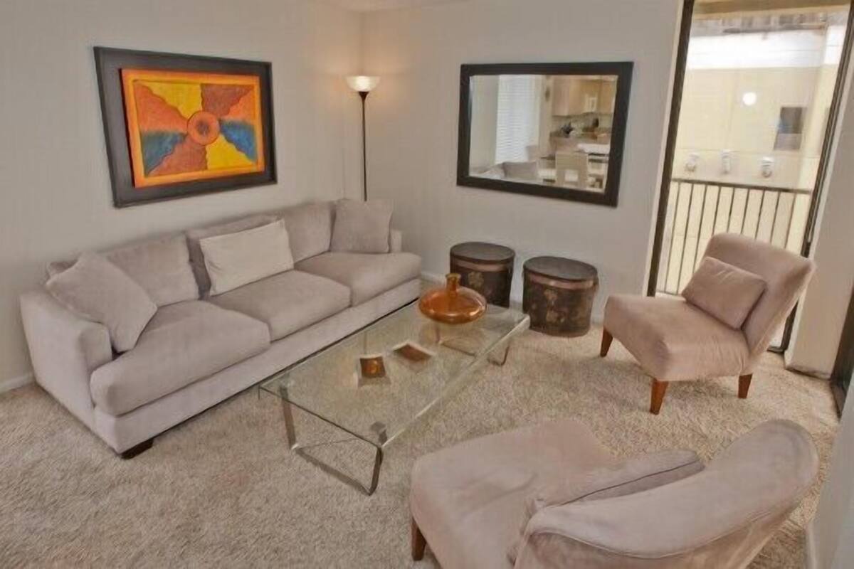 A living room with a sofa, armchair, and glass coffee table. There are two round side tables and a framed artwork on the wall.