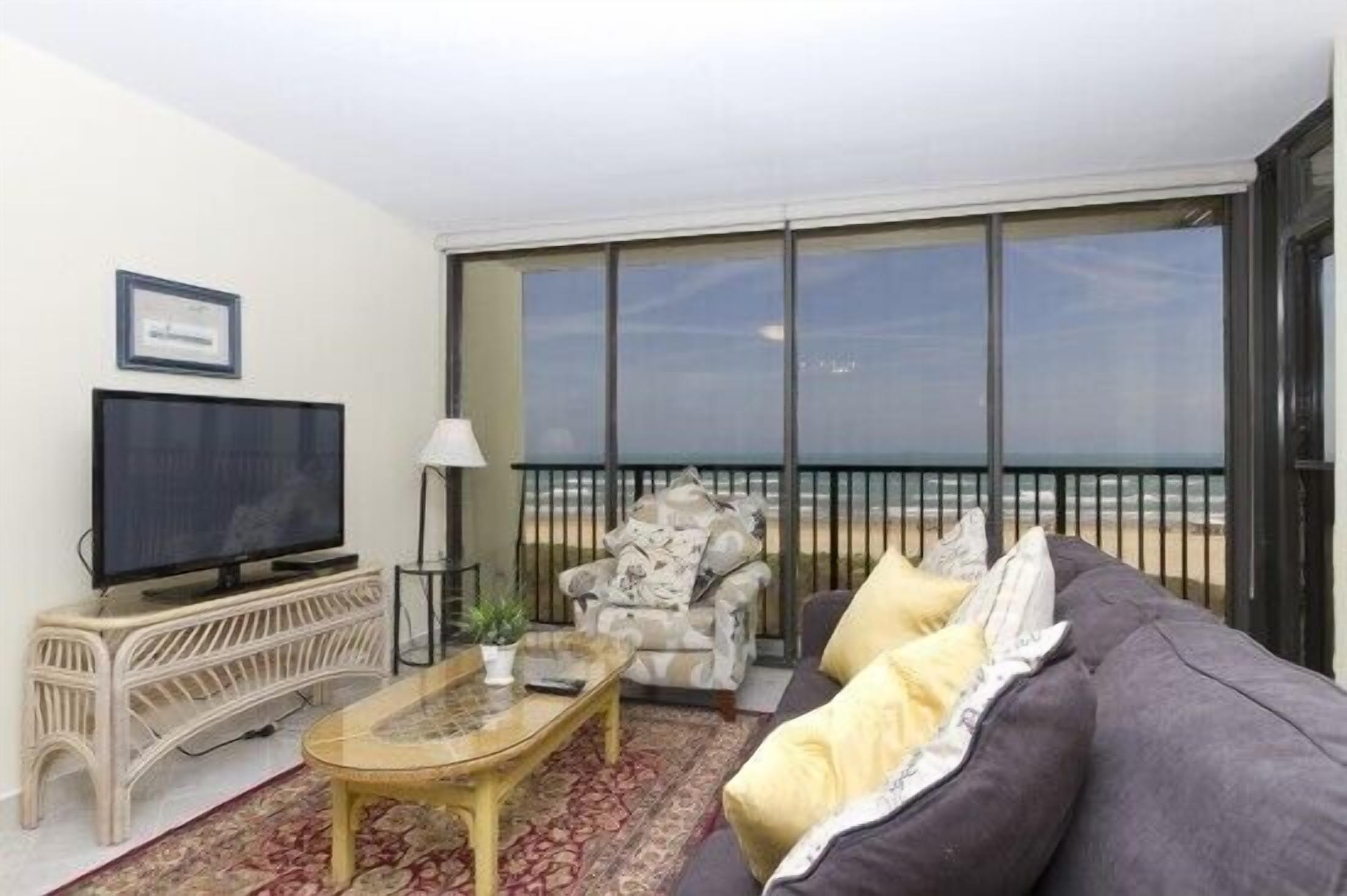 A living room with a television, a sofa, a chair, and a large window overlooking the beach.