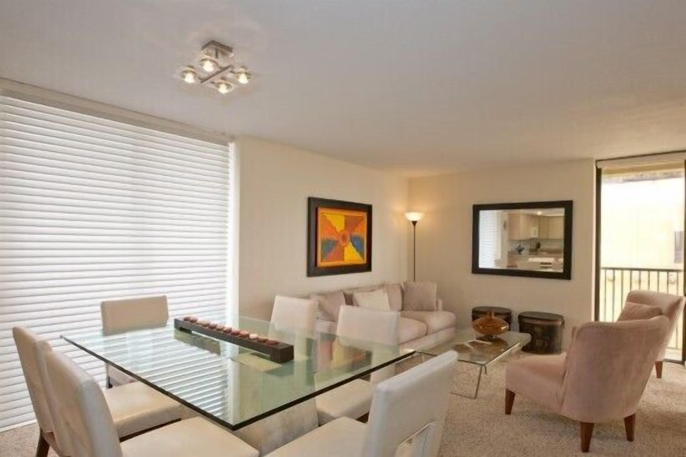 A modern living room with a glass dining table, white sofas, and a large window with blinds.