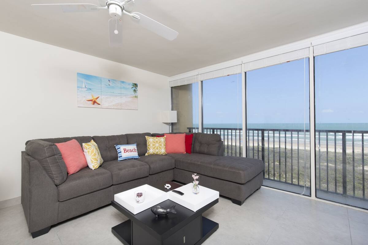 A modern living room with a grey sectional sofa, a black coffee table, and a ceiling fan. There is a large window offering a view of the beach.