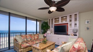 A living room with a view of the beach, a ceiling fan, a TV, and a cabinet with decorative items. - Sea Vista by South Padre Condo Rentals (South Padre Island)