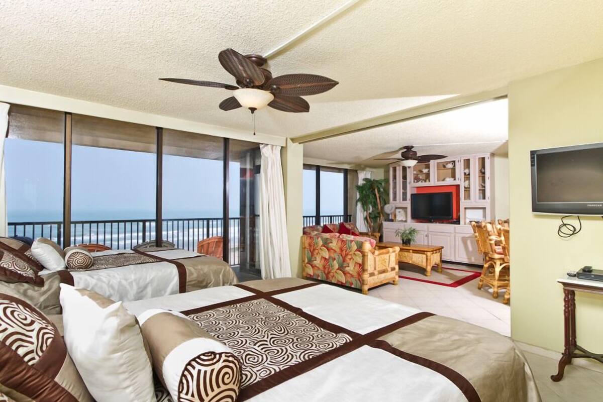 A hotel room with two beds, a balcony with a view of the ocean, a television, and a dining area.