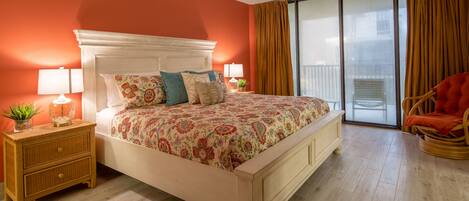 A bedroom with a large bed, a nightstand with a lamp, a potted plant, and a chair.