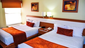 Habitación de hotel con dos camas, cada una con ropa de cama blanca y mantas color naranja. Hay dos mesitas de noche con lámparas y un teléfono. La habitación tiene una ventana con persiana marrón y dos cuadros enmarcados en la pared.