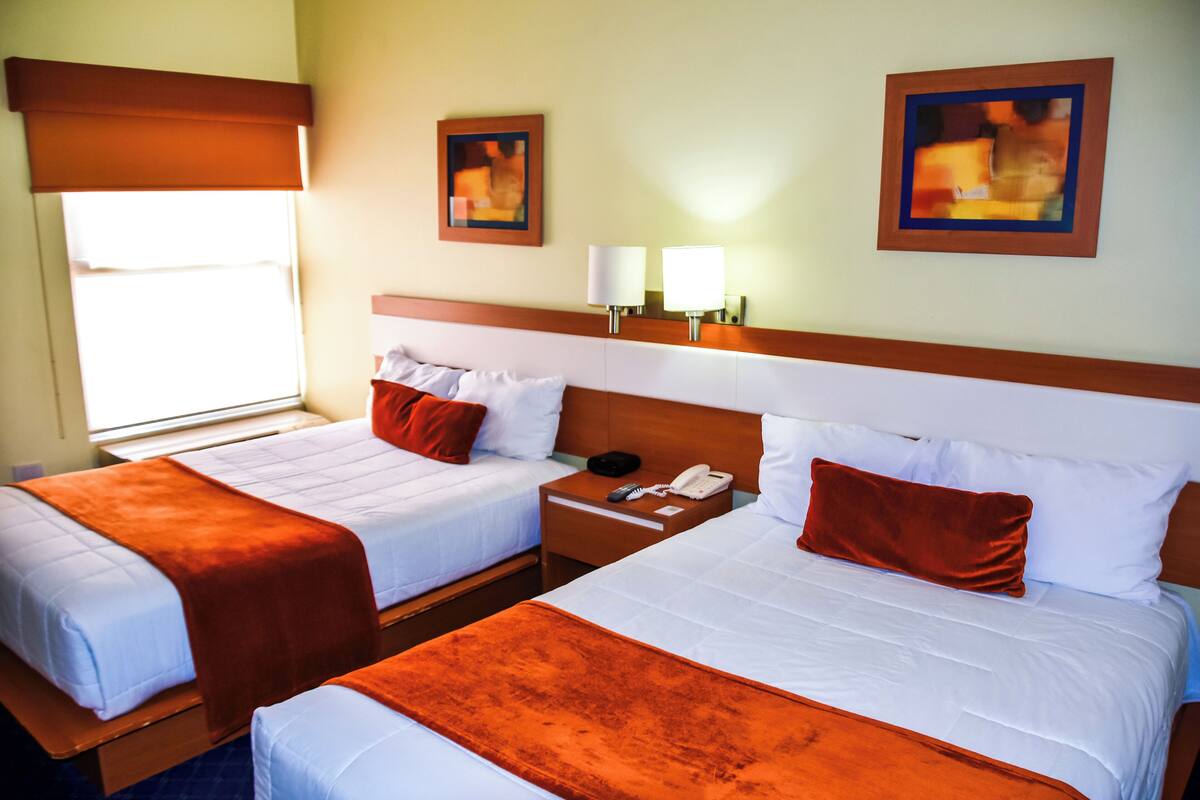 Habitación de hotel con dos camas, cada una con ropa de cama blanca y mantas color naranja. Hay dos mesitas de noche con lámparas y un teléfono. La habitación tiene una ventana con persiana marrón y dos cuadros enmarcados en la pared.
