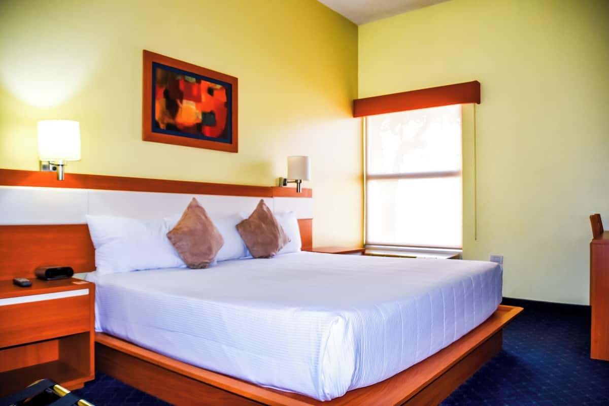 Habitación de hotel con una cama grande, cabecera de madera, dos lámparas de noche, un cuadro en la pared y una ventana con persianas.