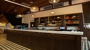 A bar area with a checkered floor, wooden bar counter, pendant lighting, and shelves with bottles and glasses. - The Chambers (George Town)
