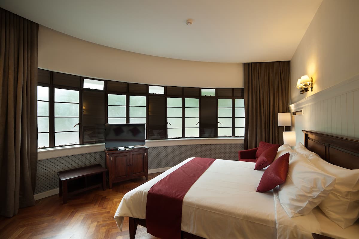 A hotel room with a large bed, a TV, a bench, and a view of the outdoors through large windows.