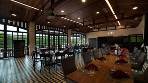 A spacious dining area with a long table set for a meal, surrounded by chairs, and large windows offering a view of greenery outside. - The Chambers (George Town)