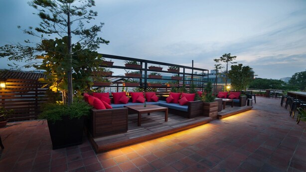 An outdoor seating area with wooden benches and red cushions, surrounded by potted plants and a metal railing. - The Chambers (George Town)
