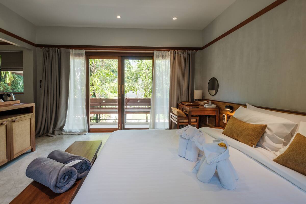 A hotel room with a large bed, a desk, a chair, a small table, and a view of the outdoors through a sliding door.