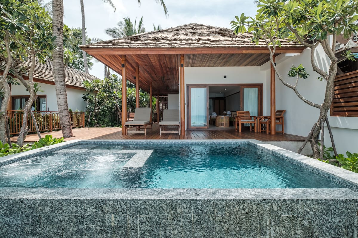 A swimming pool with a wooden deck, a covered seating area, and a building with a tiled roof.