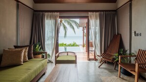 Ein Zimmer mit Holzbalkendecke, einer Couch mit Kissen, einem Holzstuhl, einem kleinen Tisch und Blick auf den Ozean durch eine Glastür.