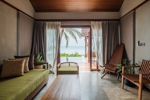 A room with a wooden ceiling, a sofa with cushions, a wooden chair, a small table, and a view of the ocean through a glass door.