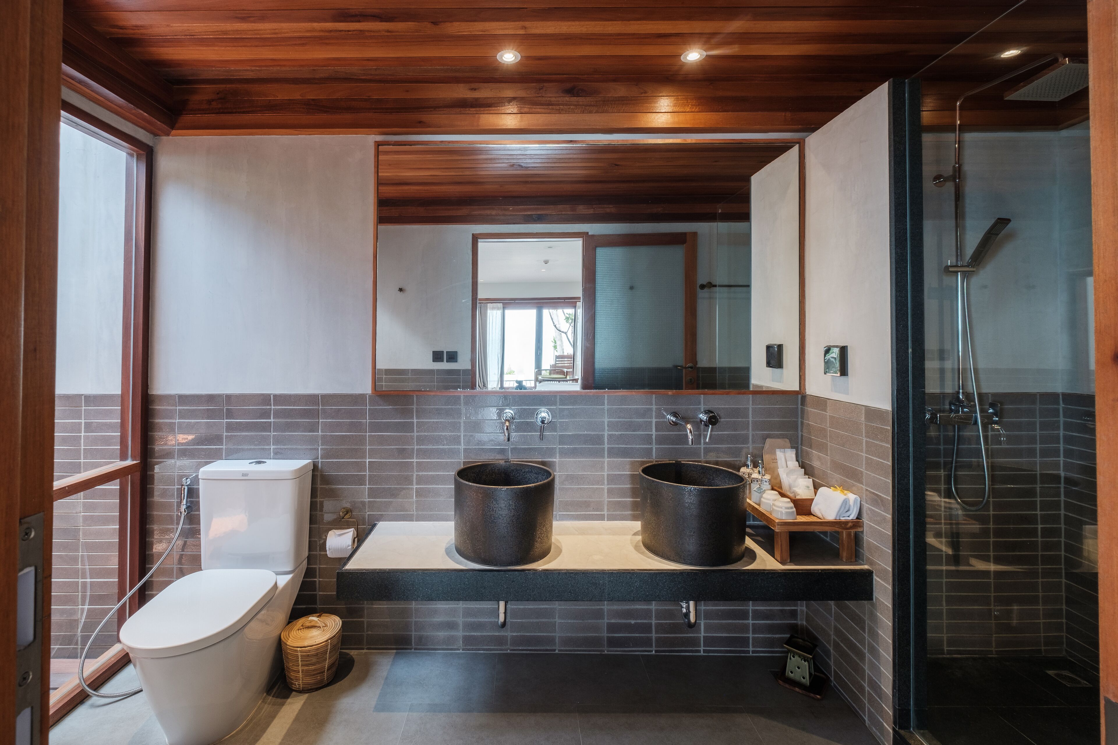 A modern bathroom with a wooden ceiling, a large mirror, two sinks, a toilet, and a glass-enclosed shower.