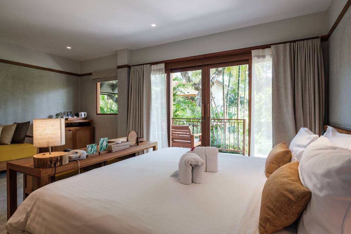 A hotel room with a large bed, a desk with a lamp, a chair, and a view of greenery outside.