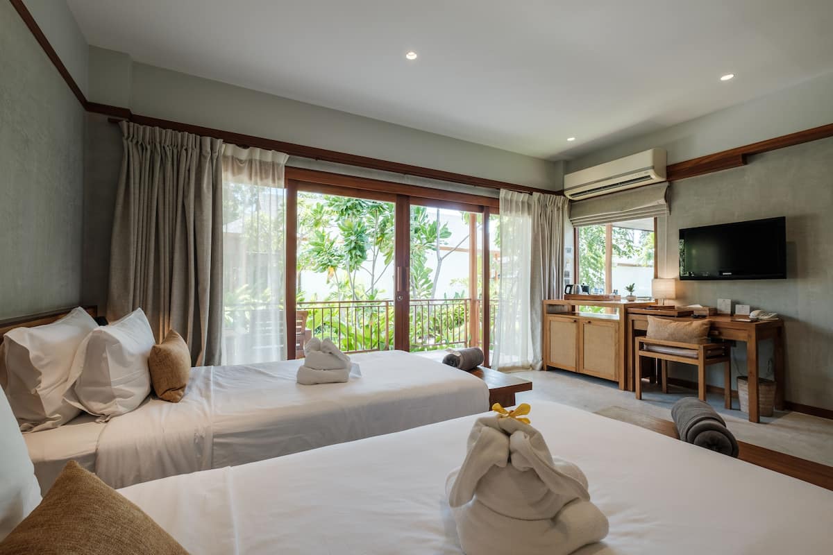 A hotel room with two beds, a desk, a TV, and a balcony with a view of greenery.