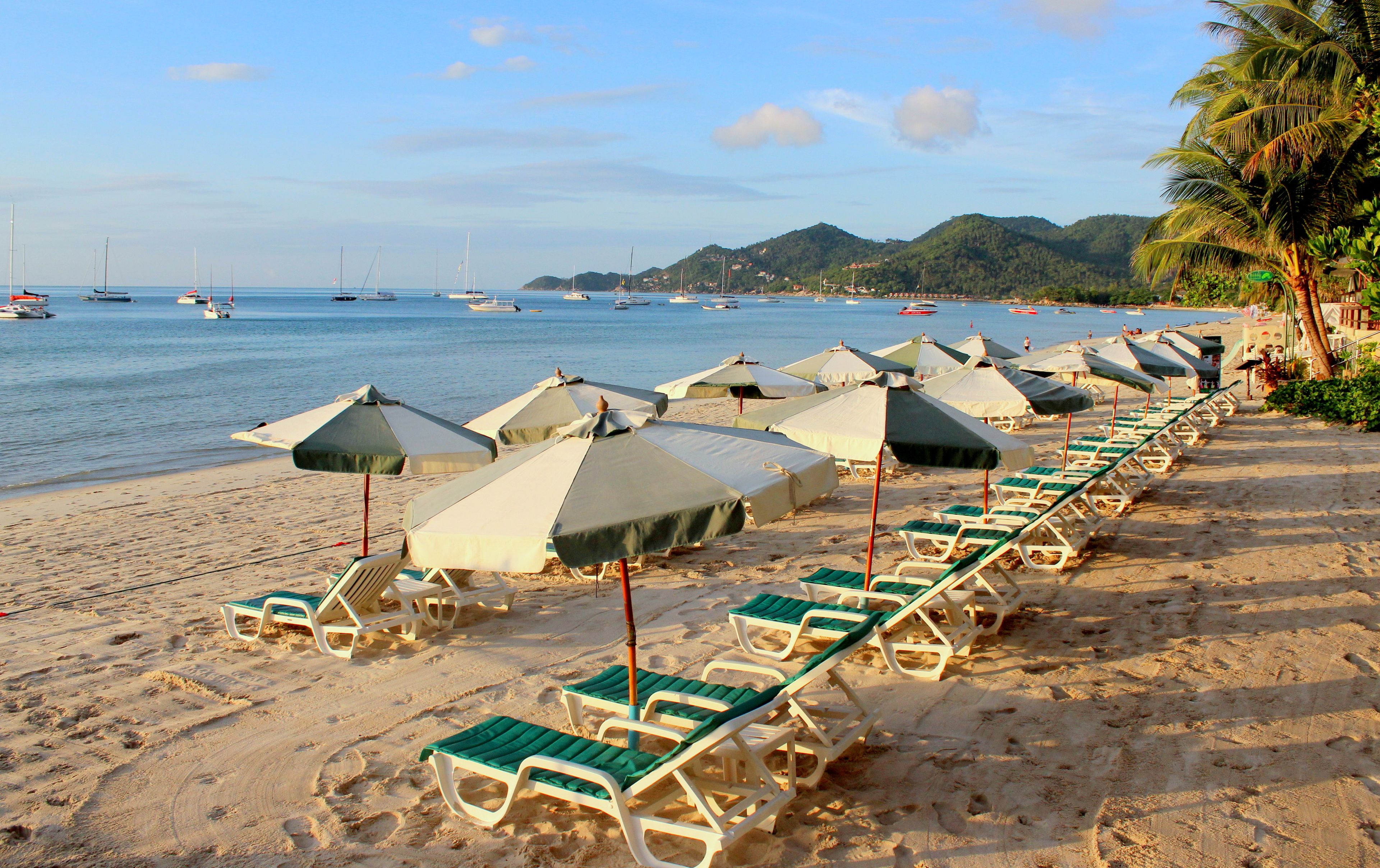 Uma praia com fileiras de espreguiçadeiras e guarda-sóis, céu claro e barcos na água.