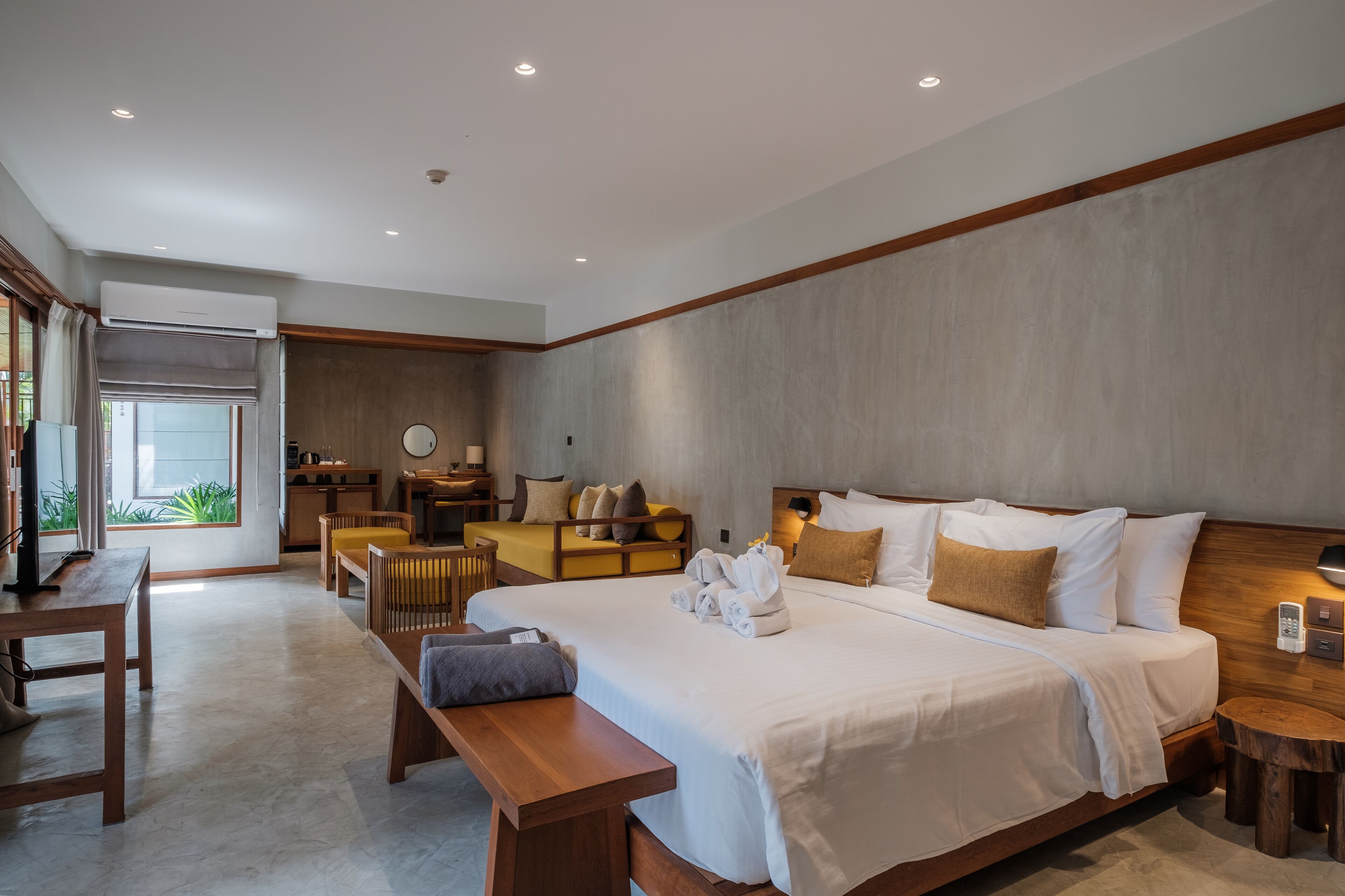 A modern hotel room with a large bed, wooden furniture, a small desk, and a view of greenery outside.