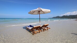 Ein Strand mit zwei Holzliegen unter einem weißen Sonnenschirm, klares blaues Wasser und sanfte Hügel in der Ferne.