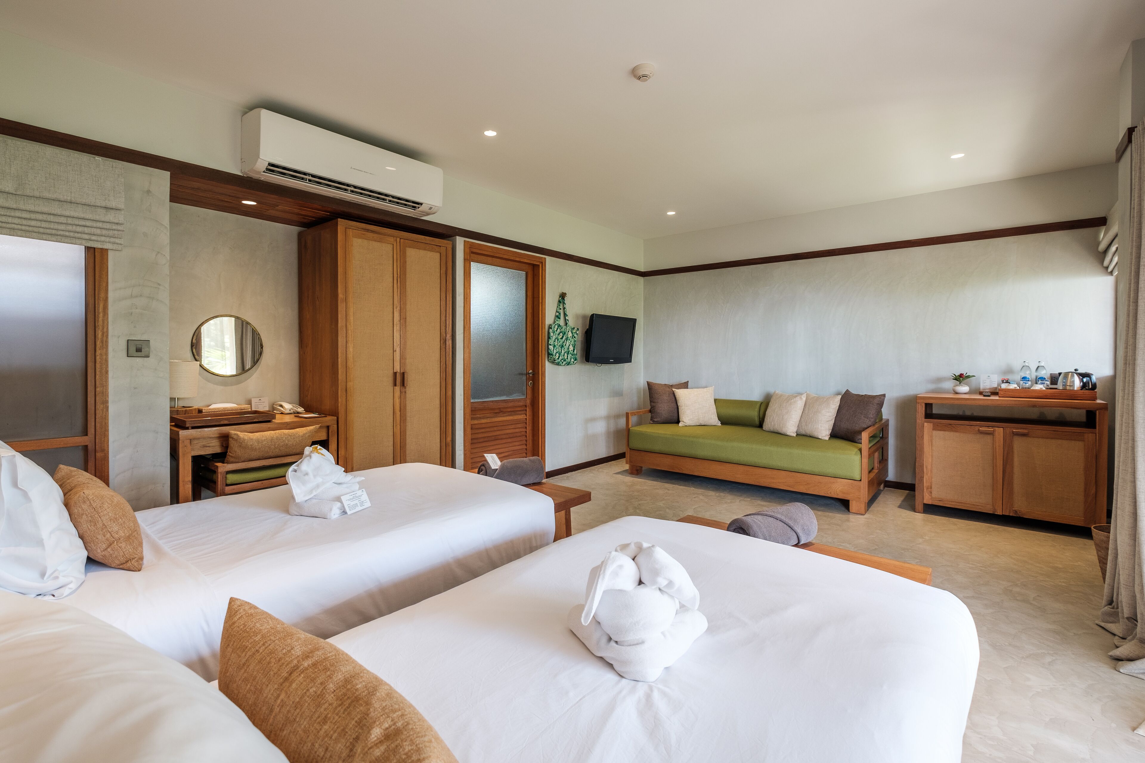 Habitación de hotel con dos camas, un sofá verde, un armario de madera, un televisor y una mesita con una planta.