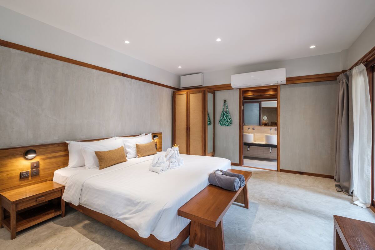 A modern hotel room with a large bed, wooden headboard, bedside tables, a bench, a bathroom with a vanity and sink, and an air conditioning unit.