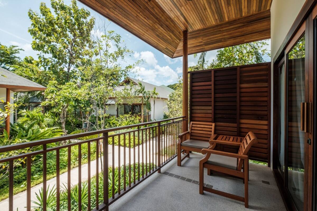 A balcony with wooden furniture, a view of a garden, and a building in the background.