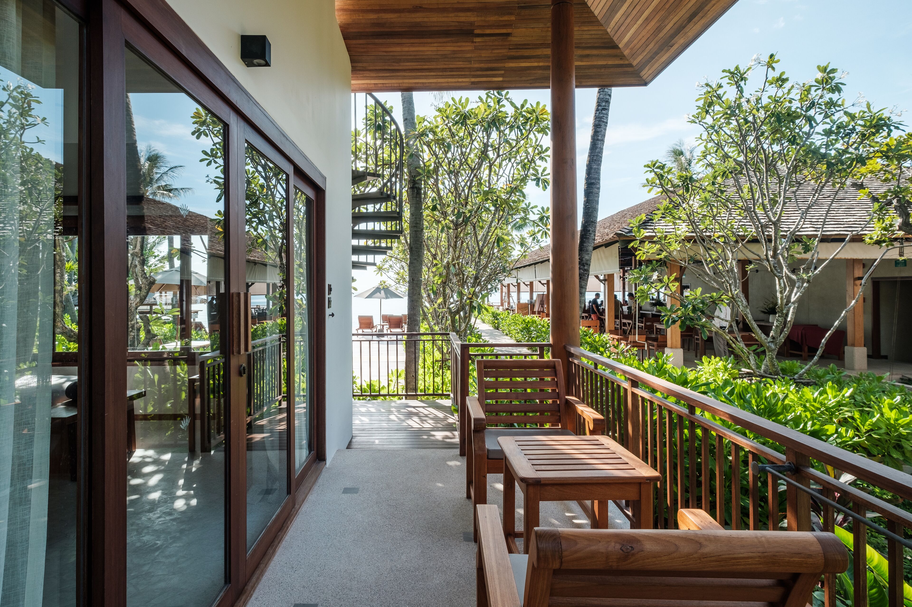 A balcony with wooden furniture, glass doors, and a view of a tropical garden and neighboring buildings.