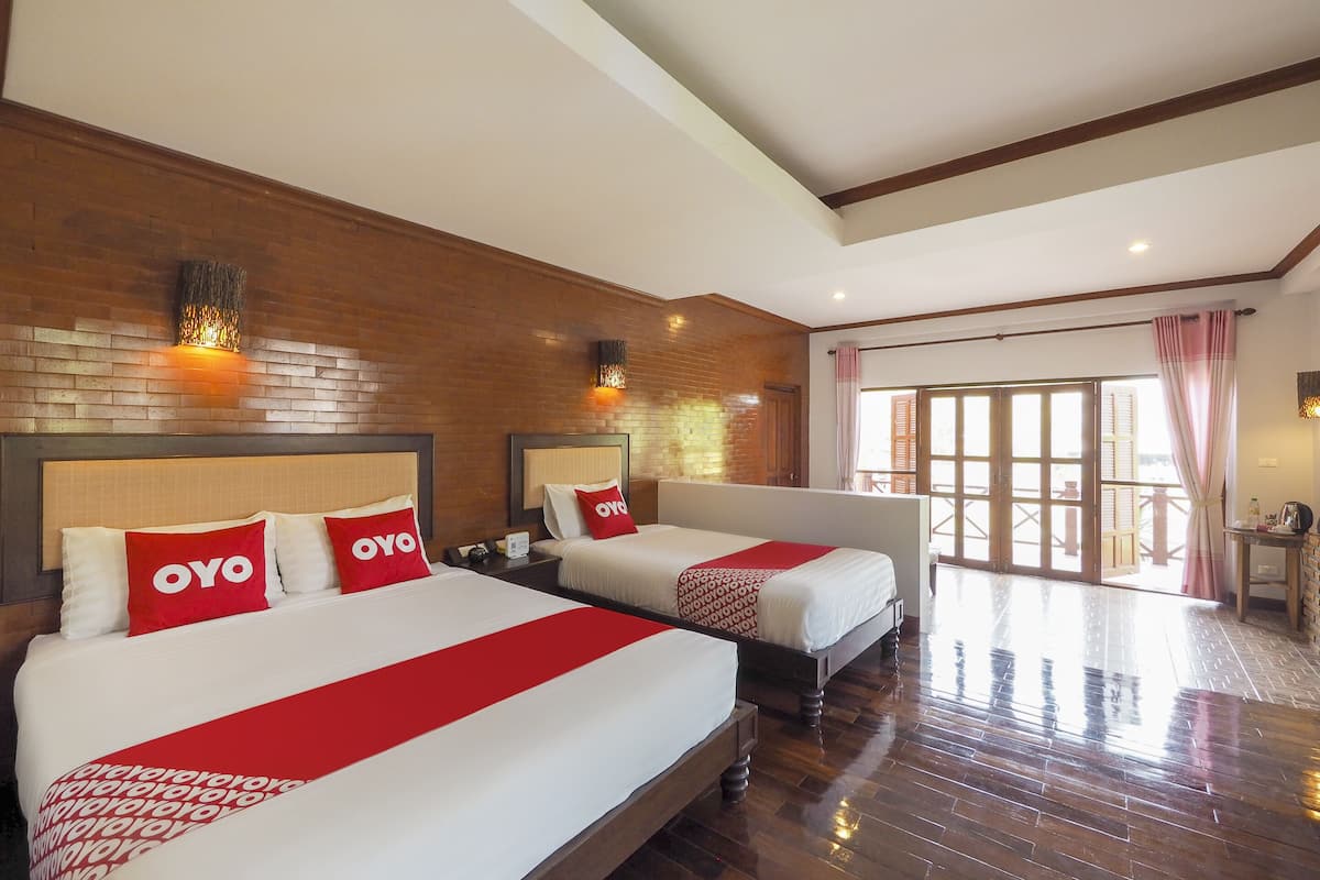 A hotel room with two beds, each with a red and white bedspread and pillows labeled "OYO". The room features wooden flooring, a brick accent wall, and a large sliding glass door leading to a balcony.