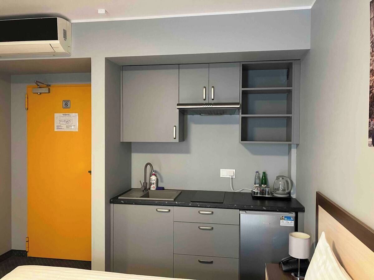 A compact hotel room with a kitchenette, a bed, a small bedside table with a lamp, and a door with a sign.