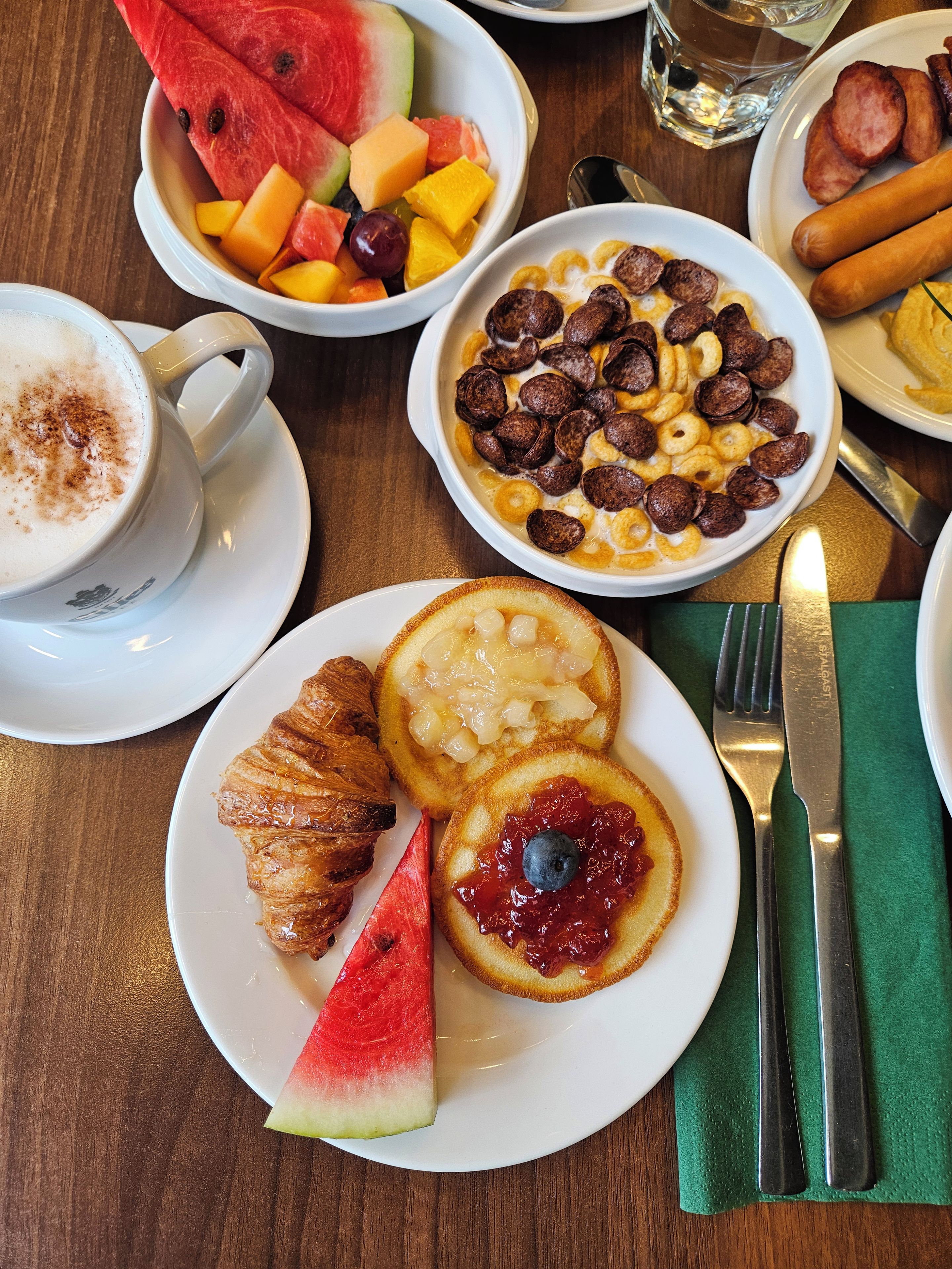 Una mesa de desayuno con diversos alimentos, como un croissant, panqueques con mermelada, un tazón de cereales y un plato con una rebanada de sandía.