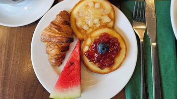 Una mesa de desayuno con diversos alimentos, como un croissant, panqueques con mermelada, un tazón de cereales y un plato con una rebanada de sandía.