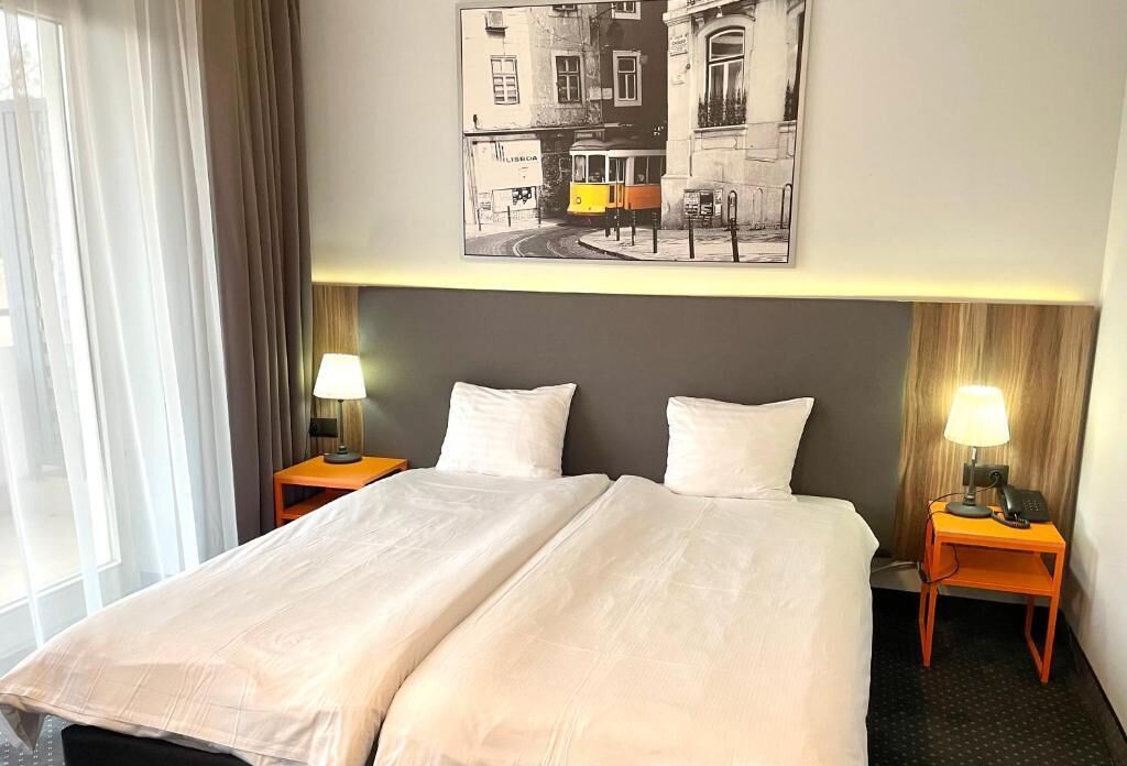 A hotel room with a double bed, two bedside tables with lamps, a framed cityscape picture on the wall, and a window with curtains.